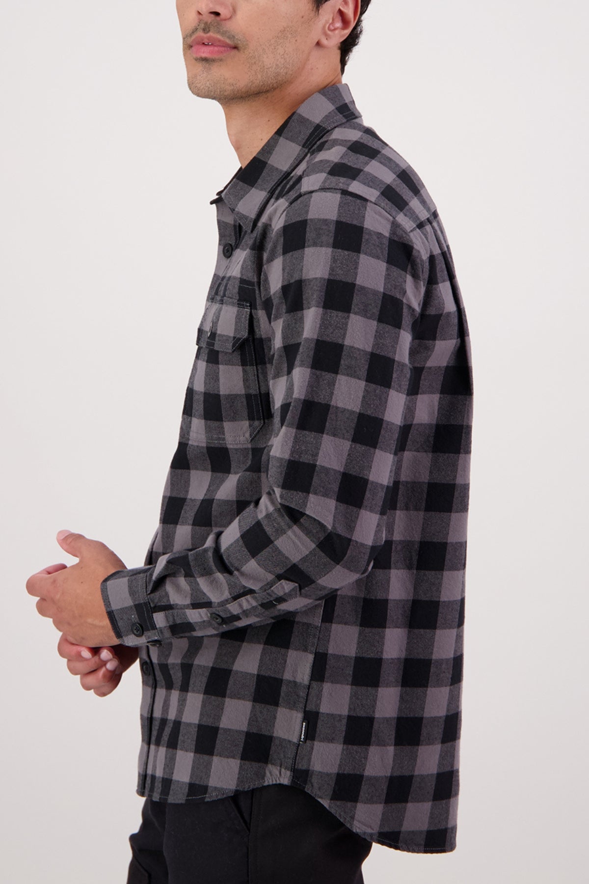 A person wearing the Swanndri Taranaki Tailor Shirt, a black and gray plaid 100% cotton men's casual shirt, with black pants stands sideways with hands loosely clasped against a plain light background.