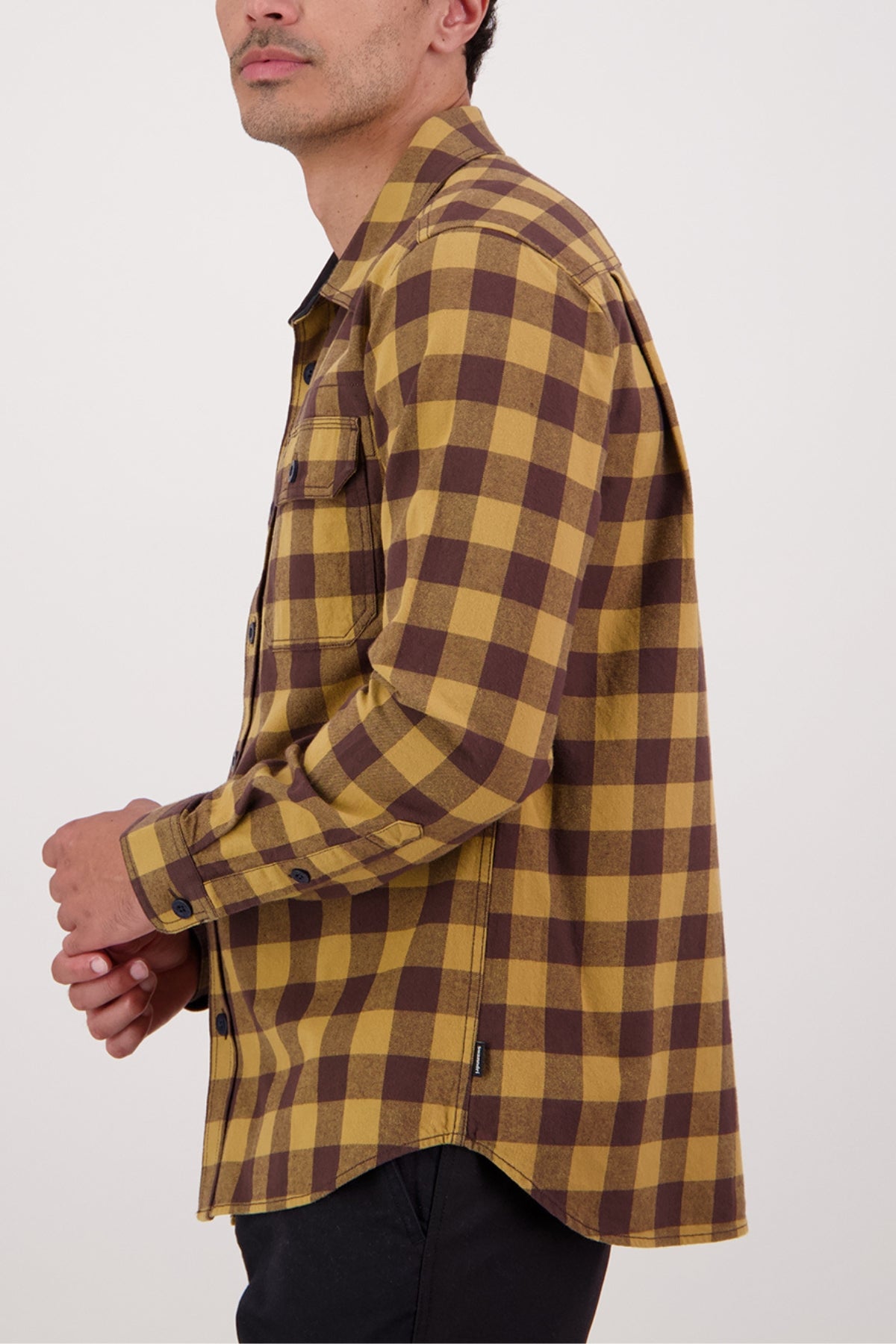 A person in the Swanndri Taranaki Tailor Shirt—long-sleeved, yellow and brown plaid 100% cotton with a chest pocket and black buttons—faces sideways against a plain white background.