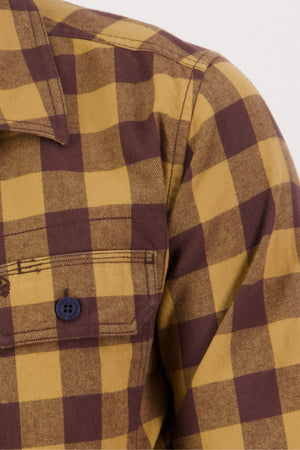Close-up of the Swanndri Taranaki Tailor Shirt, a men's 100% cotton yellow and brown plaid button-up, showing the chest pocket with black button, collar, and shoulder seam.