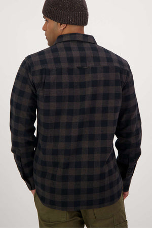 A person dressed in a Swanndri Taranaki Tailor Shirt featuring a dark checkered pattern, green pants, and a brown knit beanie is seen from behind. With their hands resting at their sides, they stand against a plain white background.
