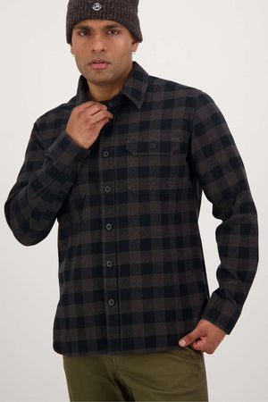 A person wearing a Swanndri Taranaki Tailor Shirt, featuring a brown and black checkered button-up design made from premium 100% cotton, and a brown knit beanie stands against a plain background. They are adjusting their shirt collar with one hand and have a neutral expression.