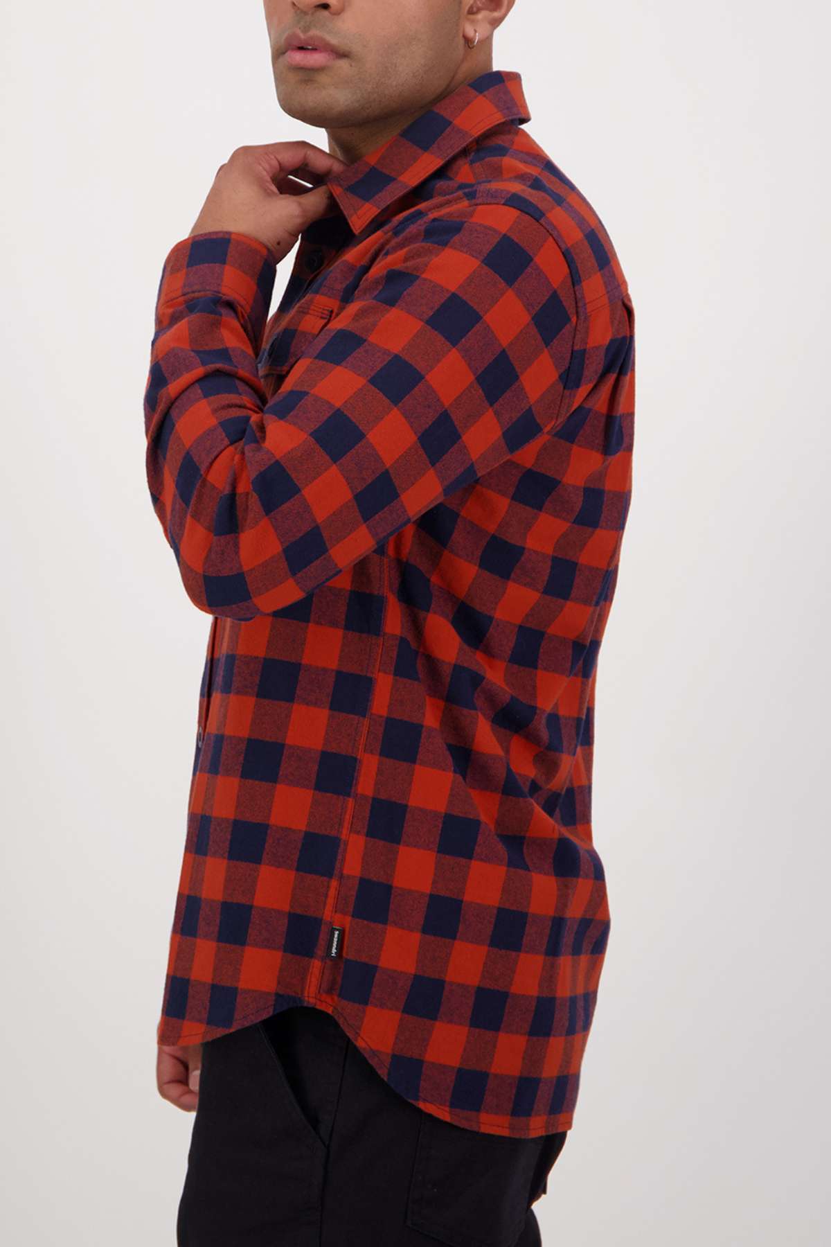 A person stands sideways, gently touching the collar of their Swanndri Taranaki Tailor Shirt in a long-sleeve, red and navy blue checkered pattern. The 100% cotton shirt features a tailored fit against a plain white background.