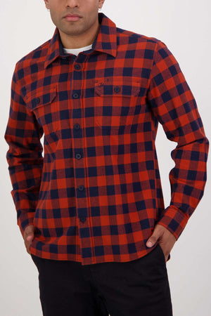 A person in a Swanndri Taranaki Tailor Shirt poses against a plain backdrop. This red and blue plaid, 100% cotton button-up boasts a collar and front pockets, its tailored fit accentuated by hands casually resting in the pockets.