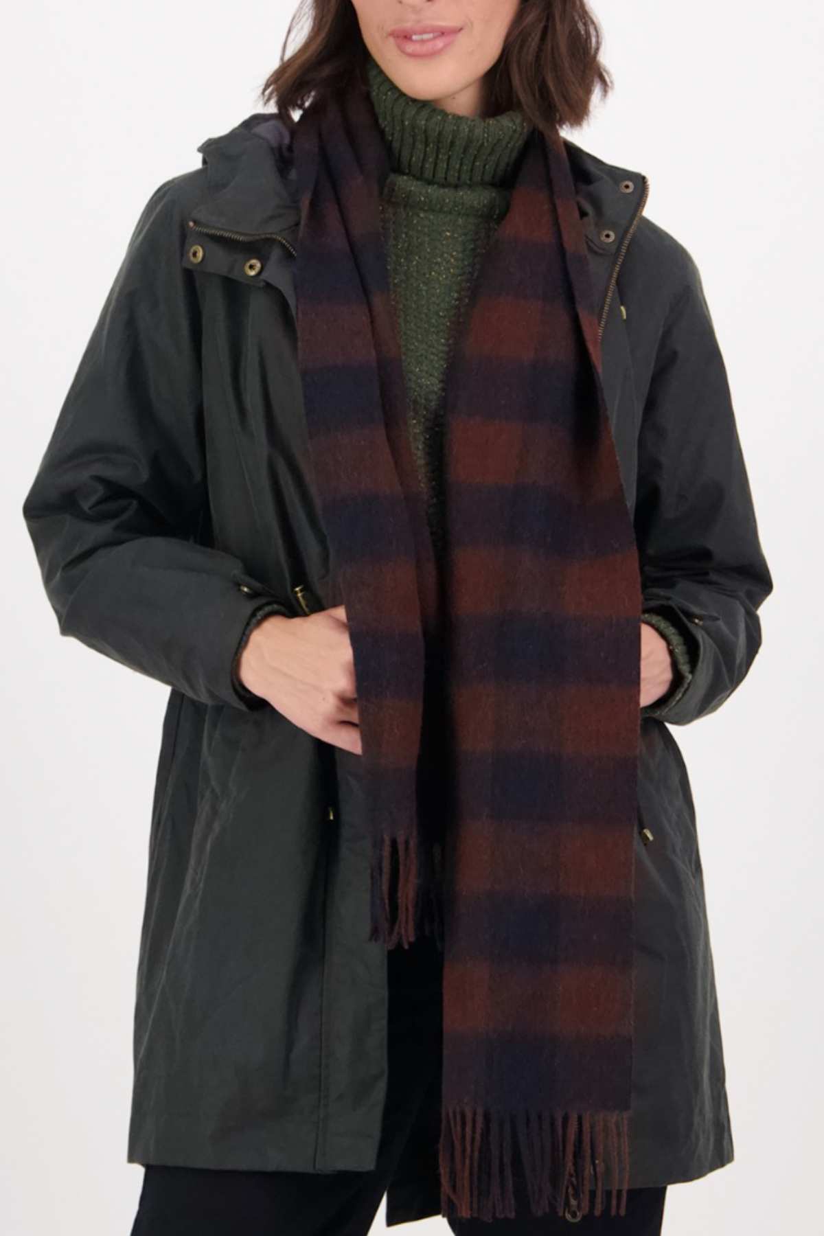 A person wearing Swanndri The Scarfie—a brown-and-navy plaid brushed wool scarf with fringed ends—along with a dark green jacket and a green turtleneck sweater, standing against a plain white background.