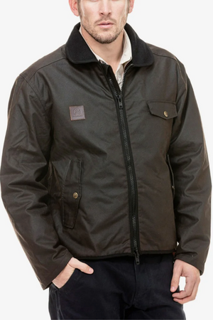 A man wearing the Swanndri Waimak Oilskin Jacket—a dark brown waxed cotton zip-up with a collar and chest and side pockets—pairs it with dark pants while standing against a plain white background.
