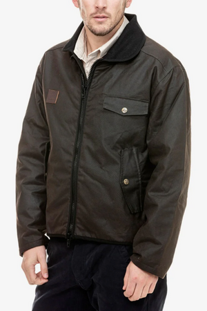 A man wears the Swanndri Waimak Oilskin Jacket, a dark brown waxed cotton zip-up with buttoned chest and side pockets, over a light collared shirt, standing against a plain white background.