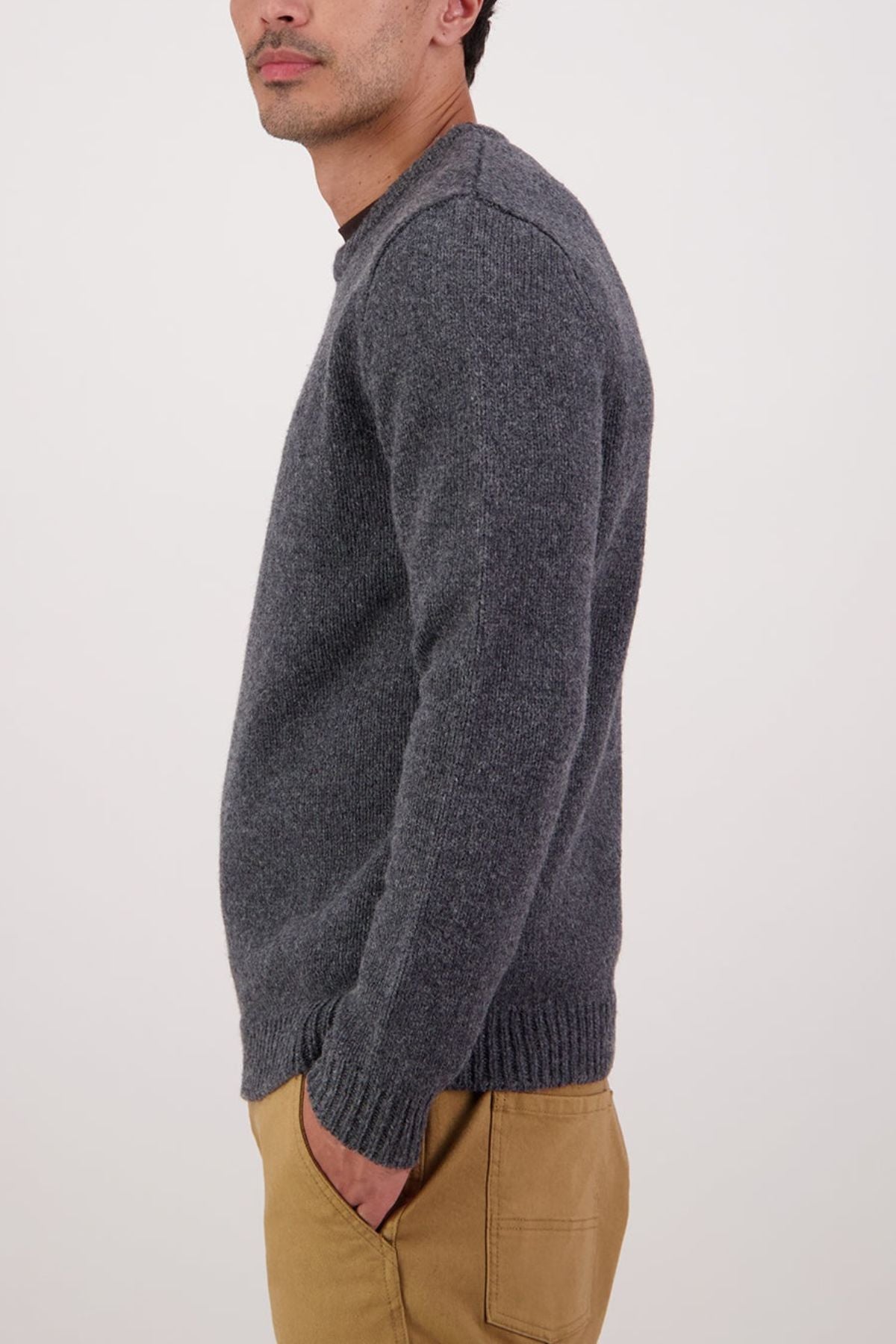 A man wearing the Swaandri Waylins Wool Knit Crew in grey and tan pants stands in profile with hands in his pockets against a plain light background.