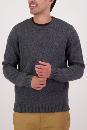 Wearing the Swaandri Waylins Wool Knit Crew in gray over a dark shirt and tan pants, a person stands against a plain light background, looking to the side with hands clasped in front.