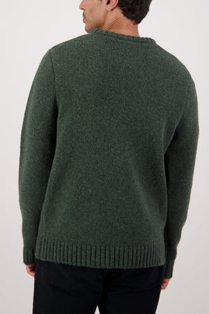 A person is seen from the back wearing a thick, dark green Swaandri Waylins Wool Knit Crew sweater and black pants, standing against a plain light background.