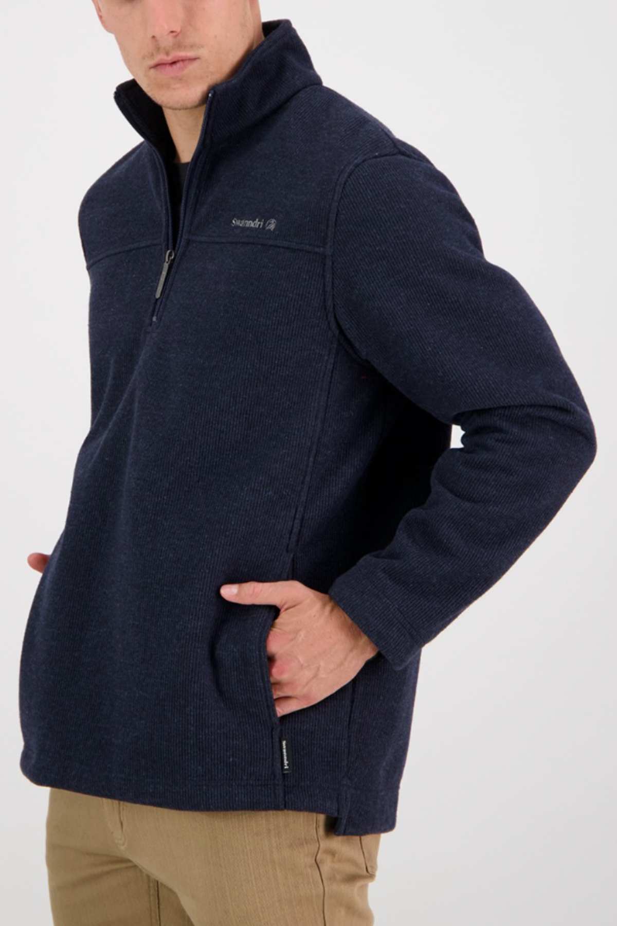 A man is wearing the Swanndri Weka Sweater, a dark blue, heavyweight quarter-zip pullover with his left hand in his pocket. This breathable and windproof sweater features a zippered chest pocket. He is also wearing tan pants and standing against a plain, light-colored background.