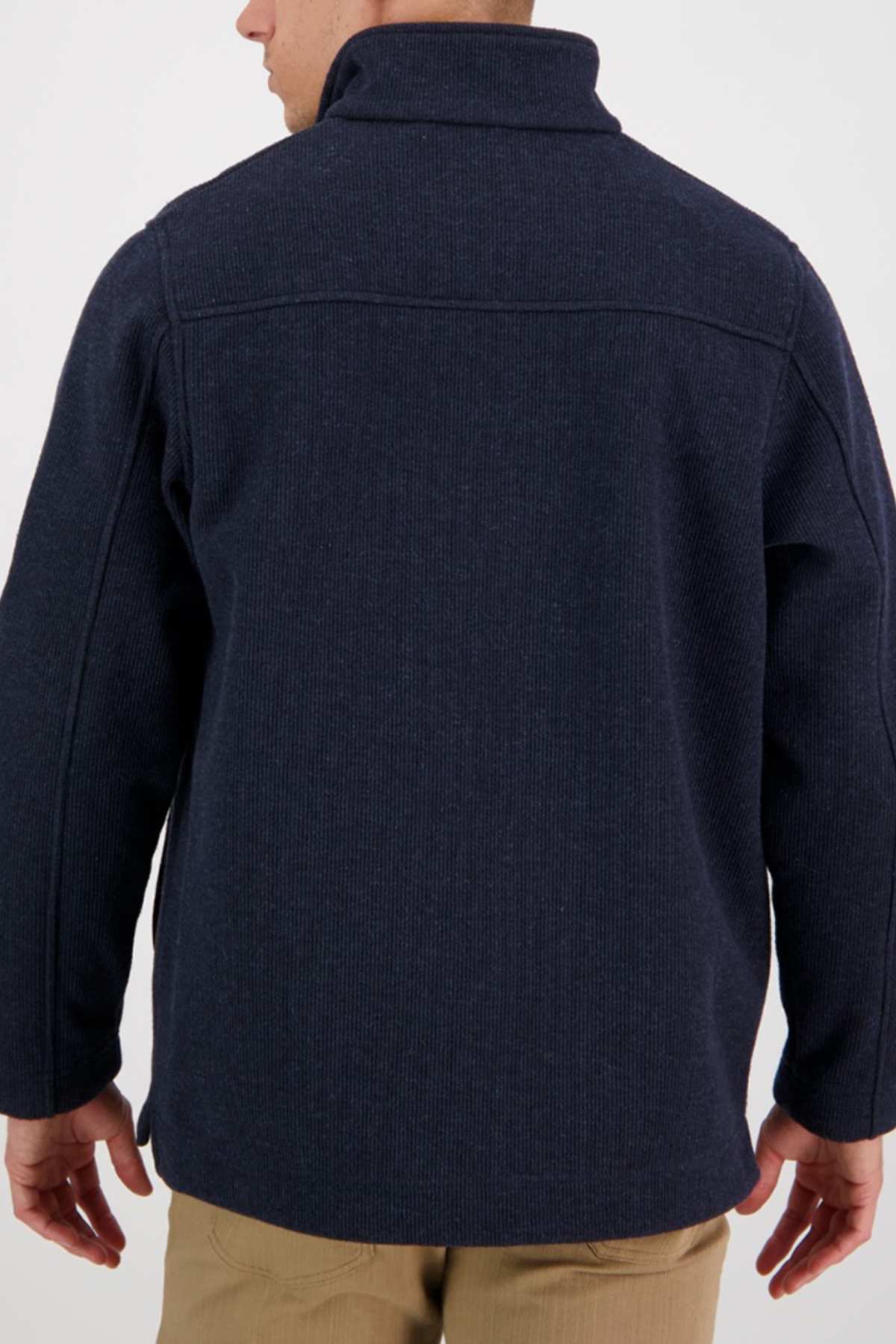 A person is seen from the back wearing a Swanndri Weka Sweater, a dark navy blue, breathable and windproof jacket with a high collar. The sweater features visible seam lines running across the shoulders and down the sleeves. Complementing the outfit, they are also wearing light brown pants. The background is plain and light-colored.