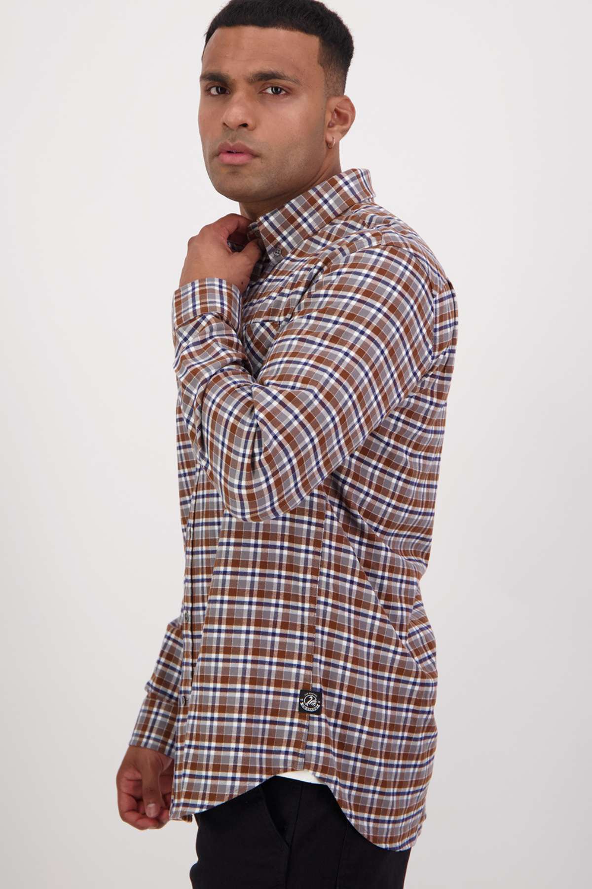 A man stands against a plain white backdrop, angled slightly to the side. He wears a classic fit Swanndri Wendon Shirt in a brown and white plaid pattern made from 100% cotton, paired with black pants. With one hand on his shirt collar, he looks directly at the camera.