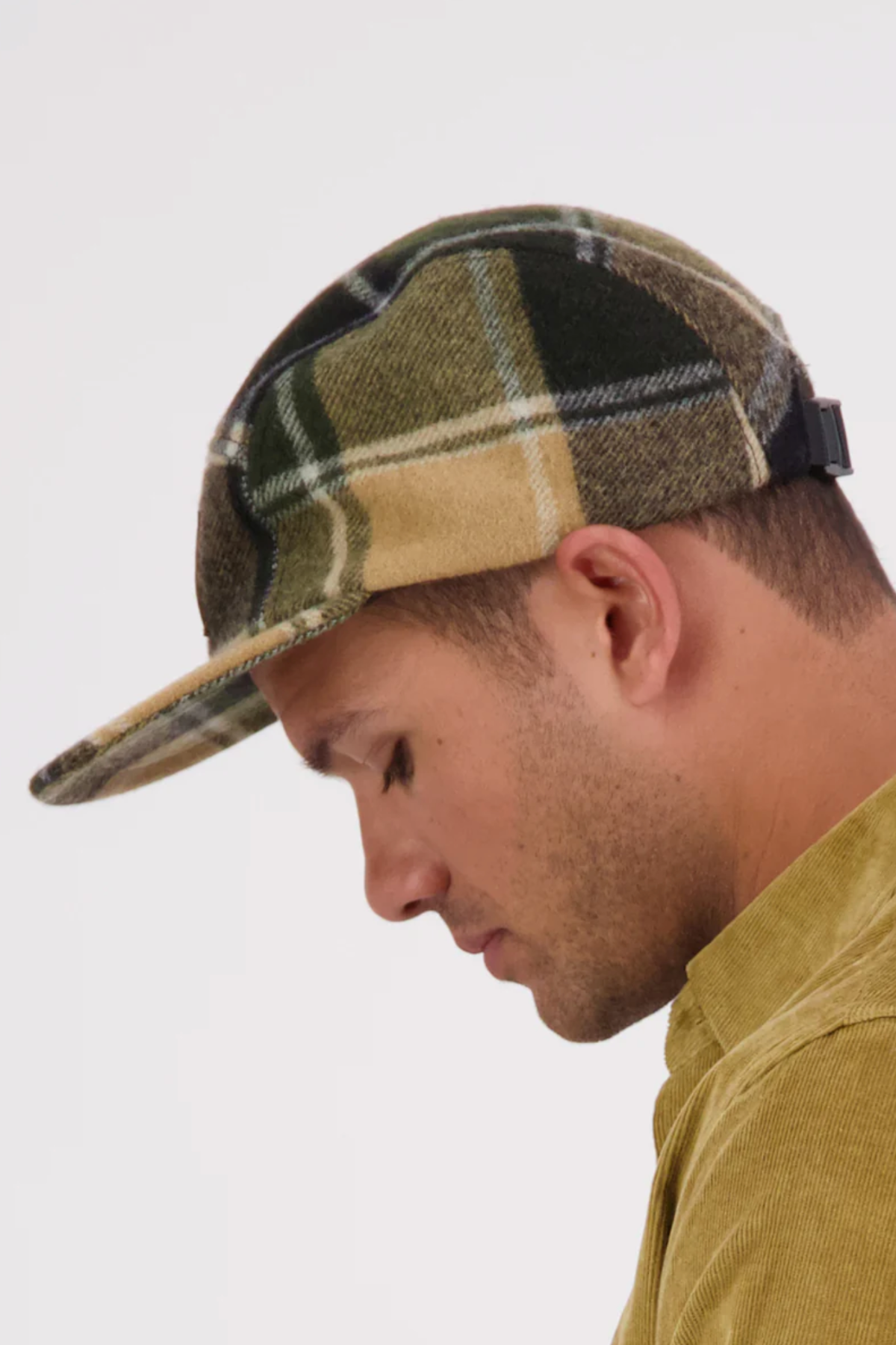 A man in a tan corduroy shirt looks down, wearing a Swanndri Wentworth Valley v2 Wool Cap with a flat brim and adjustable buckle, set against a plain white background.