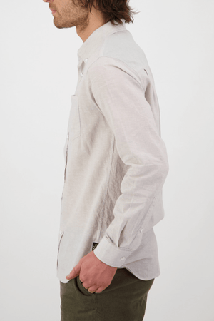 A person with long hair is clad in a light grey tailored-fit Swanndri Wolcott v2 Long Sleeve Shirt and olive green pants. They are facing sideways, showcasing the outfit's profile against a plain white background.