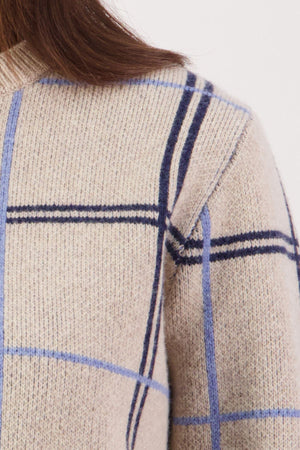 Close-up of the Swanndri Yasmin Check Womens Knit in beige merino wool with blue and navy plaid. A portion of a woman's shoulder, upper arm, and brown hair is seen against a plain light background.
