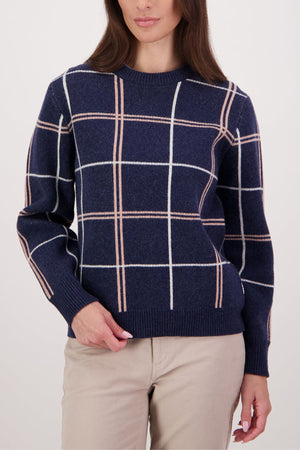 A woman stands against a light background wearing the Swanndri Yasmin Check Womens Knit in navy blue with white and light pink windowpane plaid, paired with beige pants.