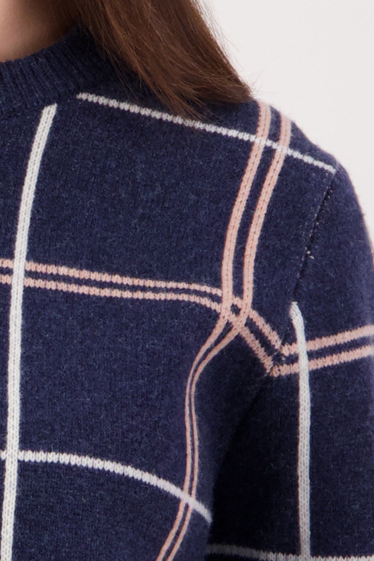 Close-up of the Swanndri Yasmin Check Womens Knit sweater in navy blue, featuring a white and pink windowpane plaid pattern. The image shows the shoulder and upper chest, with some brown hair visible.