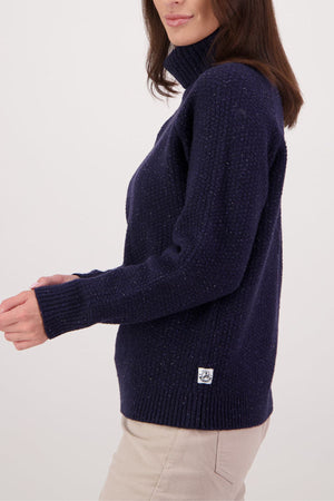 A woman shown from the side wears a navy Swanndri Lyttelton v2 Roll Neck Knit jumper with a small white tag by the hem and light beige pants, standing against a plain background.