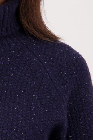 Close-up of a person wearing the Swanndri Lyttelton v2 Roll Neck Knit in textured dark blue lambswool. Only the shoulder, neck, and some brown hair are visible against a plain background.