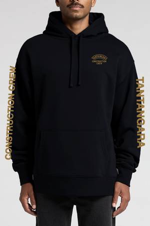 A man wears the Tantangara Construction Crew Hoodie PRE-ORDER in black with gold lettering on the chest and sleeves. This hoodie features a front pocket, drawstrings, and is available for pre-order.
