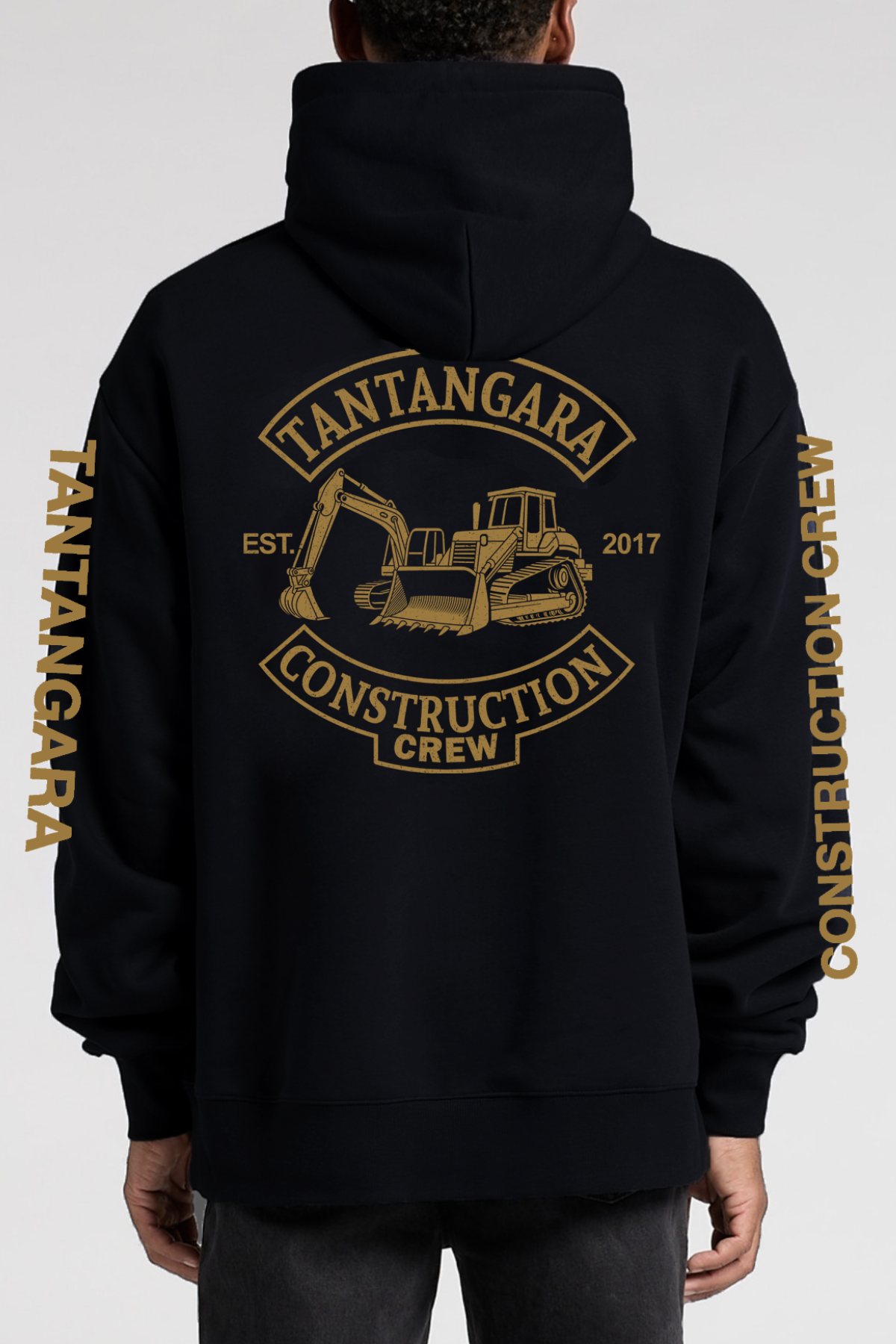 A man wears the Tantangara Construction Crew Hoodie PRE-ORDER in black with gold lettering on the chest and sleeves. This hoodie features a front pocket, drawstrings, and is available for pre-order.