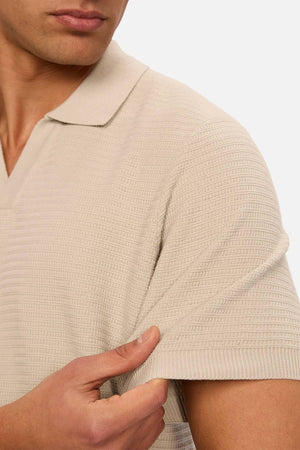 A man wearing Industrie The Alicante Polo in light beige gently pulls on its short sleeve, highlighting the men's knitted polo’s stretch and texture. The image is cropped to show only his lower face, torso, and hand.