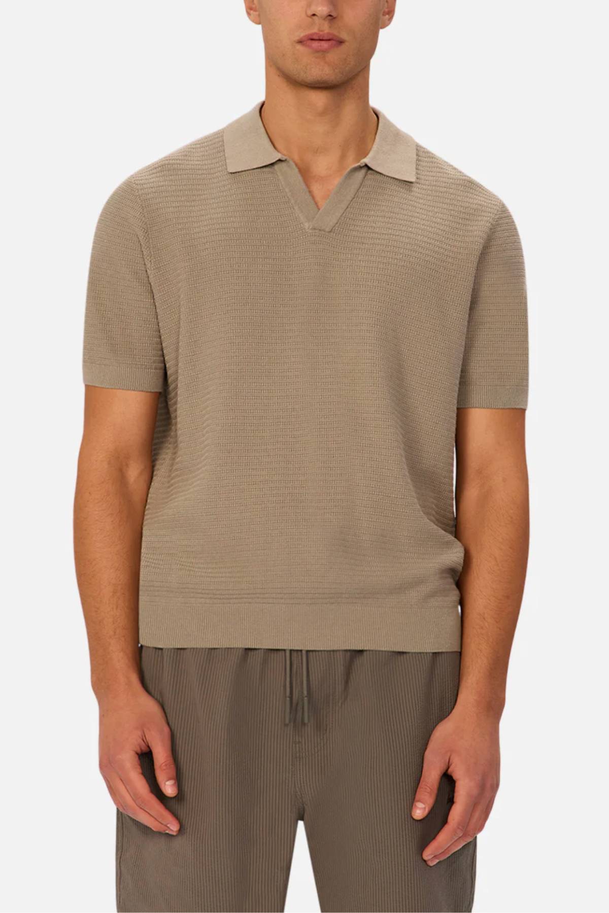 A man wears the Industrie The Alicante Polo, a short-sleeve, light brown men's cotton polo with a collar and V-neck, paired with relaxed pinstriped brown shorts, standing against a plain white background.