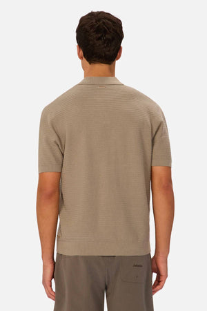 A man is shown from behind wearing the Industrie The Alicante Polo, a short-sleeved, light brown knit top, paired with dark pinstriped pants against a plain, light background.