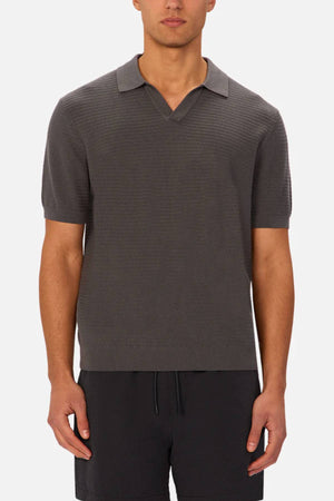 A person wears the Industrie The Alicante Polo, a short-sleeved, dark gray men's cotton polo with a V-neck collar, paired with matching dark gray shorts, standing against a light background.