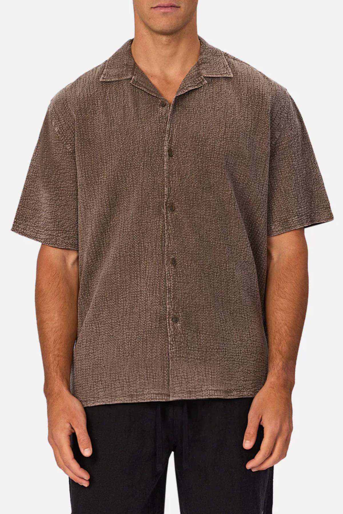 A man stands against a white background wearing the Industrie The Canyon Short Sleeve Shirt in textured brown with a camp collar, hands relaxed at his sides, paired with classic black pants.