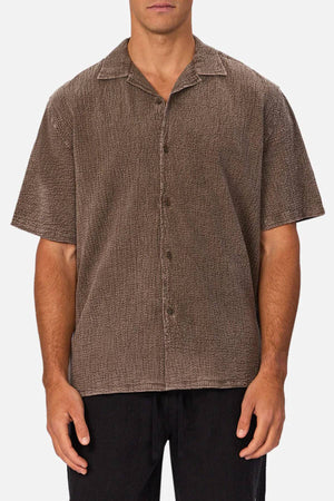 A man stands against a white background wearing the Industrie The Canyon Short Sleeve Shirt in textured brown with a camp collar, hands relaxed at his sides, paired with classic black pants.