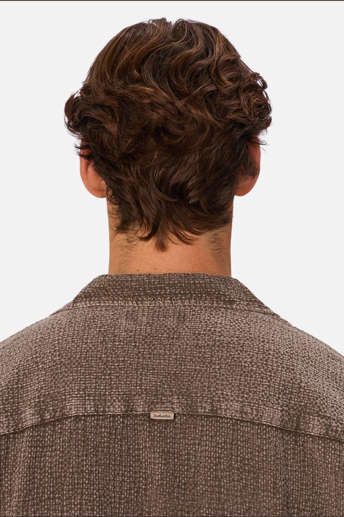 A person with short, wavy brown hair is shown from behind wearing the Industrie The Canyon Short Sleeve Shirt in textured brown, featuring a small metallic tag on the upper back. The background is plain and light-colored.