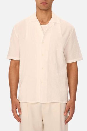 A man stands against a plain light background, wearing the Industrie The Canyon Short Sleeve Shirt with matching off-white pants. His face is partially out of frame.