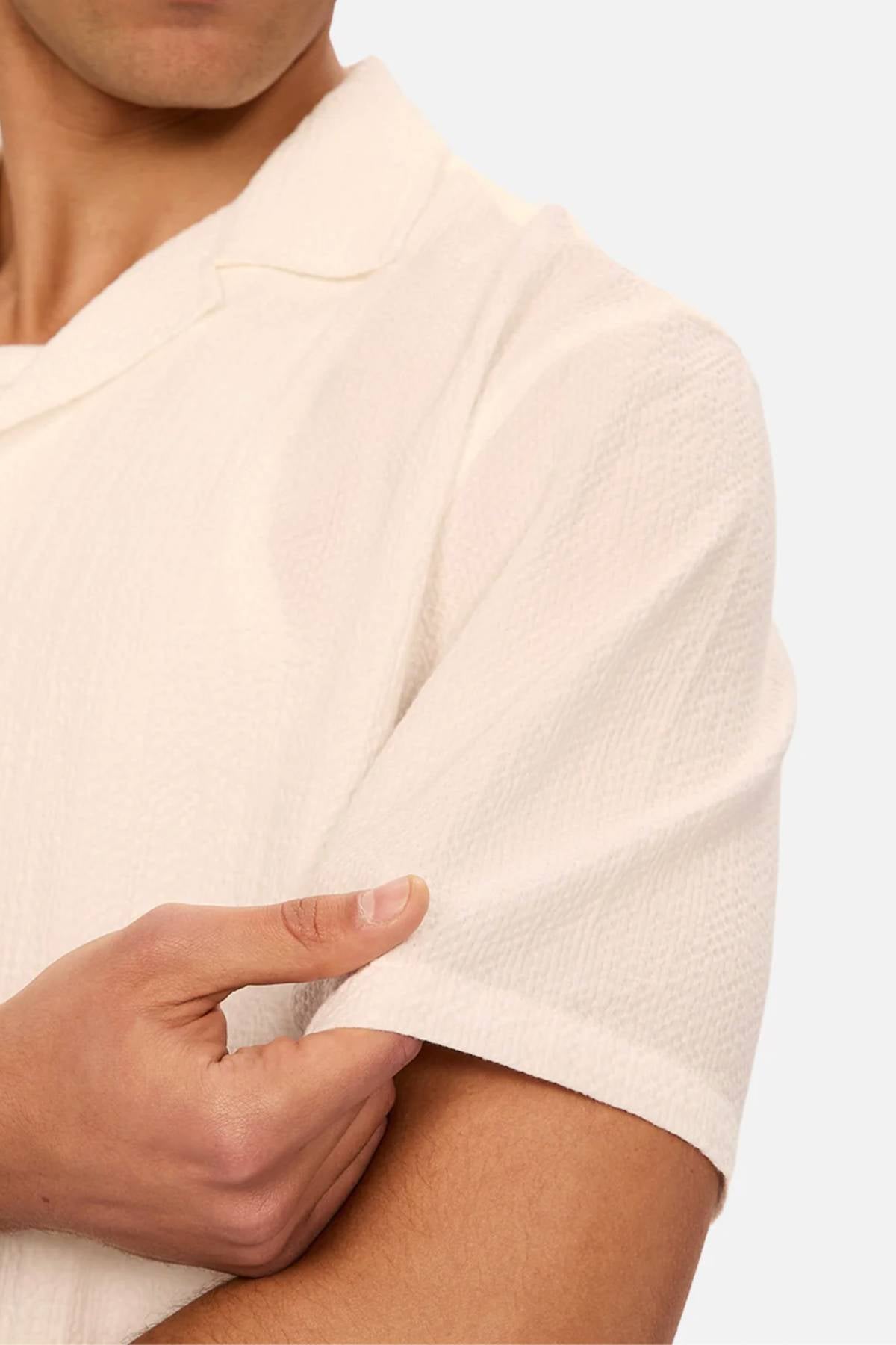 A person wearing the Industrie The Canyon Short Sleeve Shirt in off-white gently pulls at the sleeve with their right hand, highlighting the shirt's lightweight texture and fit on the upper arm.