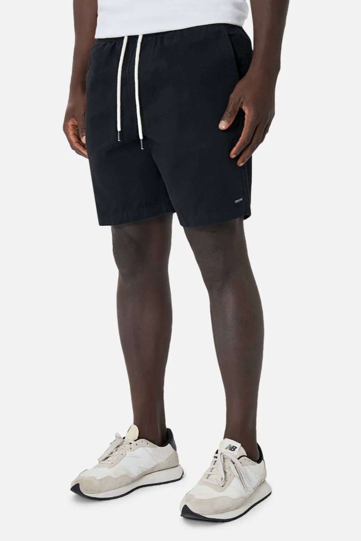 A person models Industrie The Dublin Short in black with white sneakers against a plain white background, hands relaxed at their sides. Only the lower half of their body is shown.