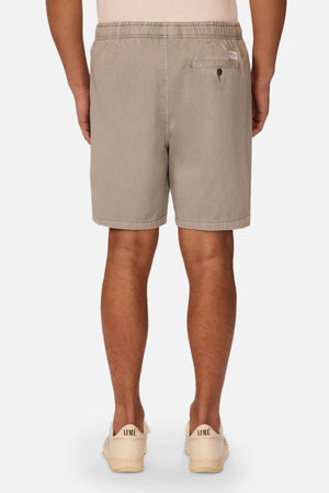 A person stands facing away, wearing Industrie The Dublin Short in light brown cotton-linen with an elastic waistband and single back pocket, paired with sneakers labeled "AIME," all against a plain white background.