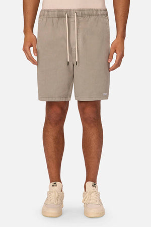 A person wears Industrie The Dublin Short, beige cotton-linen men's summer shorts with side pockets and a relaxed fit above the knees, paired with cream sneakers, standing against a plain white background.