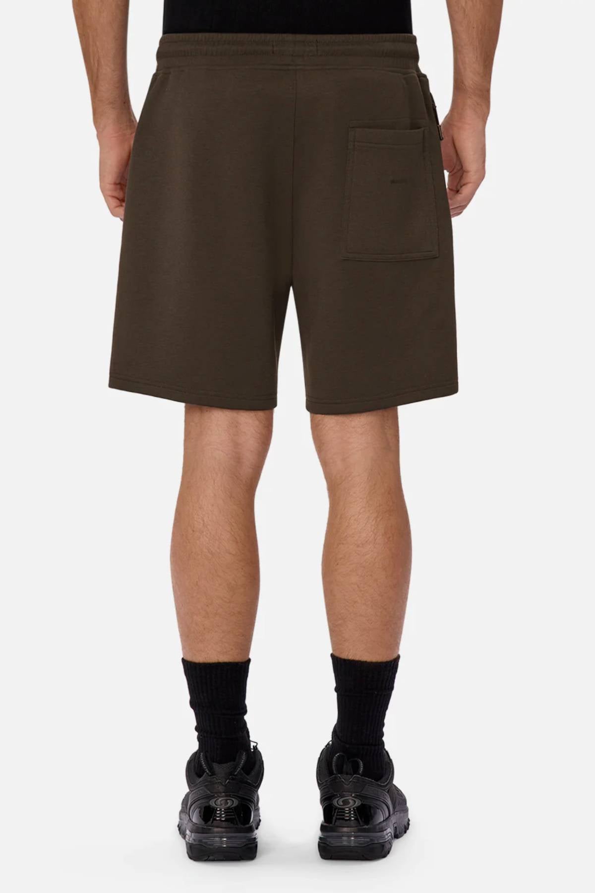 A person faces away, wearing Industrie The Tech Track Short in dark brown with a single back pocket. The versatile, comfortable shorts reach just above the knees and are paired with black socks and sneakers against a plain light background.