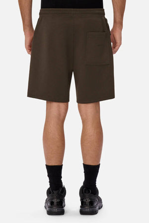 A person faces away, wearing Industrie The Tech Track Short in dark brown with a single back pocket. The versatile, comfortable shorts reach just above the knees and are paired with black socks and sneakers against a plain light background.