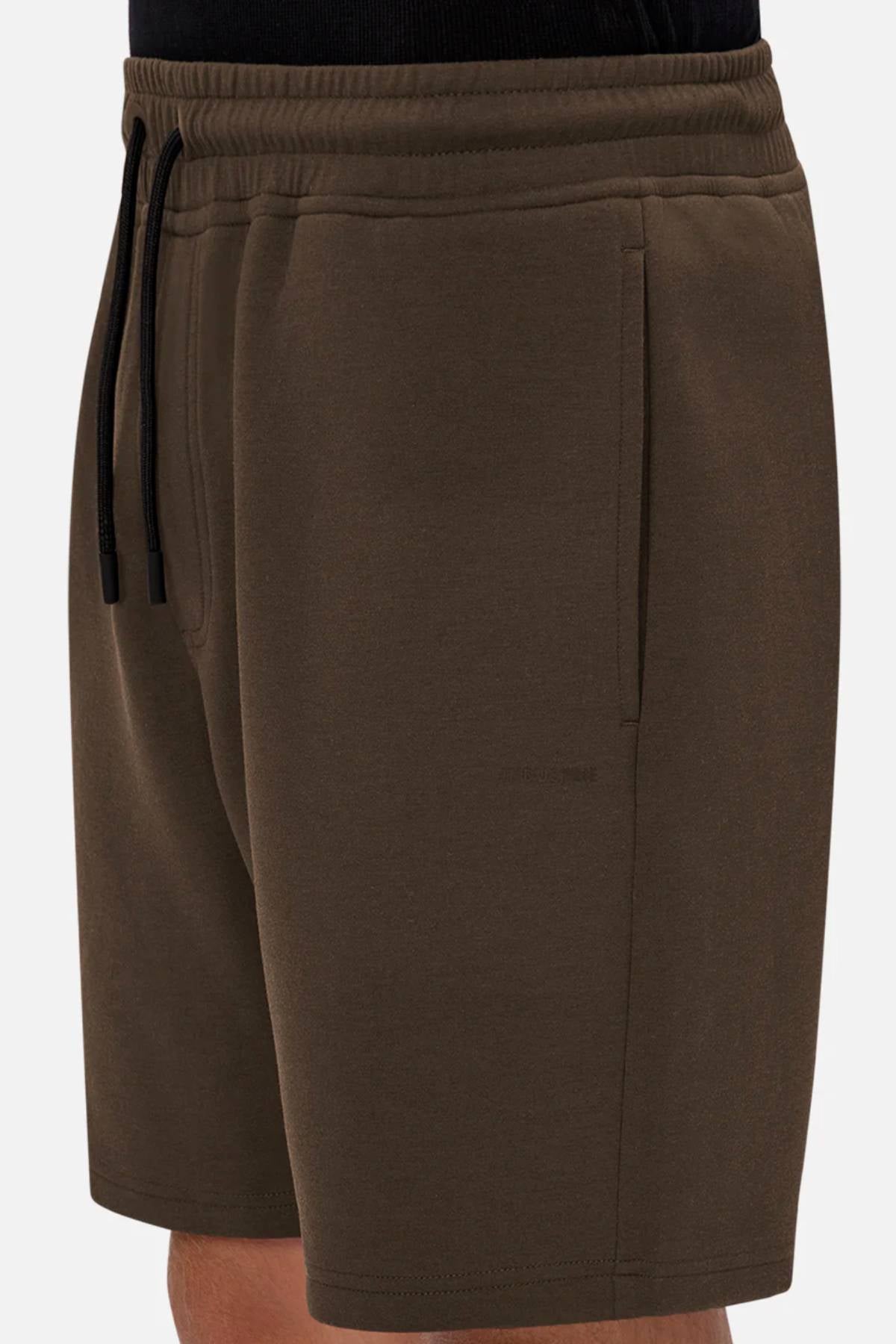 A close-up of a person wearing Industrie The Tech Track Short in dark brown, featuring an elastic waistband with black drawstrings and side pockets. The soft, textured fabric and versatile design are highlighted. Torso and one leg are partially visible.