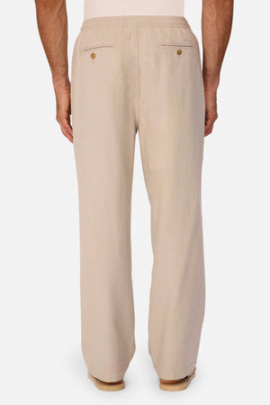 A man is shown from the back wearing Industrie The Mallorca Pant in beige, featuring an elastic waistband, pleated design, and two buttoned back pockets, standing against a white background.