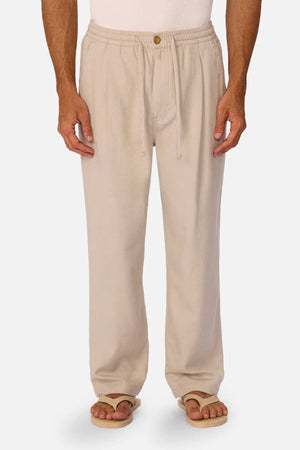 A person wearing loose-fitting beige Industrie The Mallorca Pant with a pleated design, paired with sandals and a white top, stands against a plain light background.