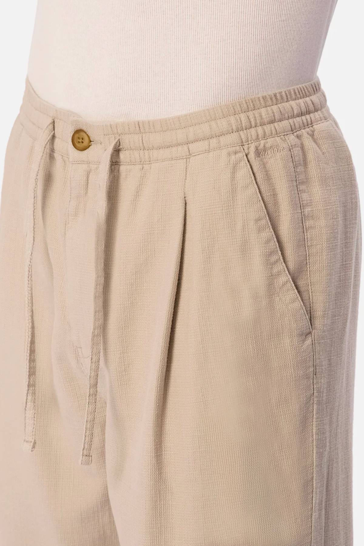 Close-up of a person wearing Industrie The Mallorca Pant in beige with a textured fabric, elastic waistband, drawstring, pleated front, side pocket, and brown button. Styled with a white ribbed top against a plain light background.