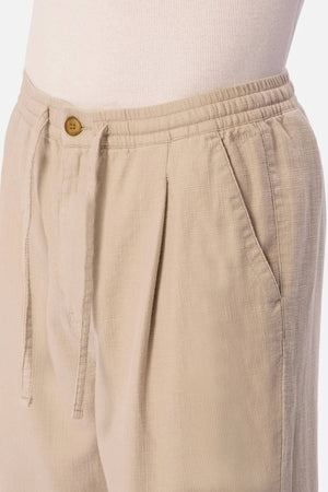 Close-up of a person wearing Industrie The Mallorca Pant in beige with a textured fabric, elastic waistband, drawstring, pleated front, side pocket, and brown button. Styled with a white ribbed top against a plain light background.