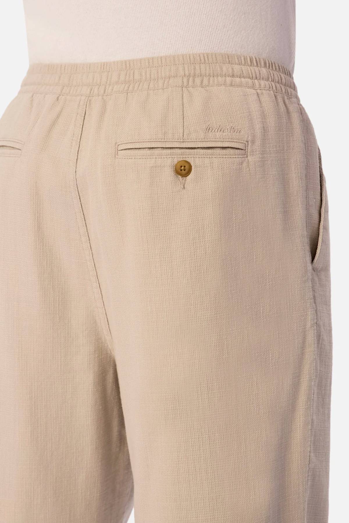 Close-up of a person wearing Industrie The Mallorca Pant in light beige, featuring breathable fabric, an elastic waistband, side pocket, and a buttoned back pocket. Styled with a tucked-in white shirt.