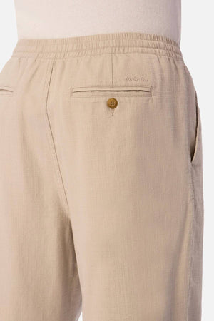 Close-up of a person wearing Industrie The Mallorca Pant in light beige, featuring breathable fabric, an elastic waistband, side pocket, and a buttoned back pocket. Styled with a tucked-in white shirt.