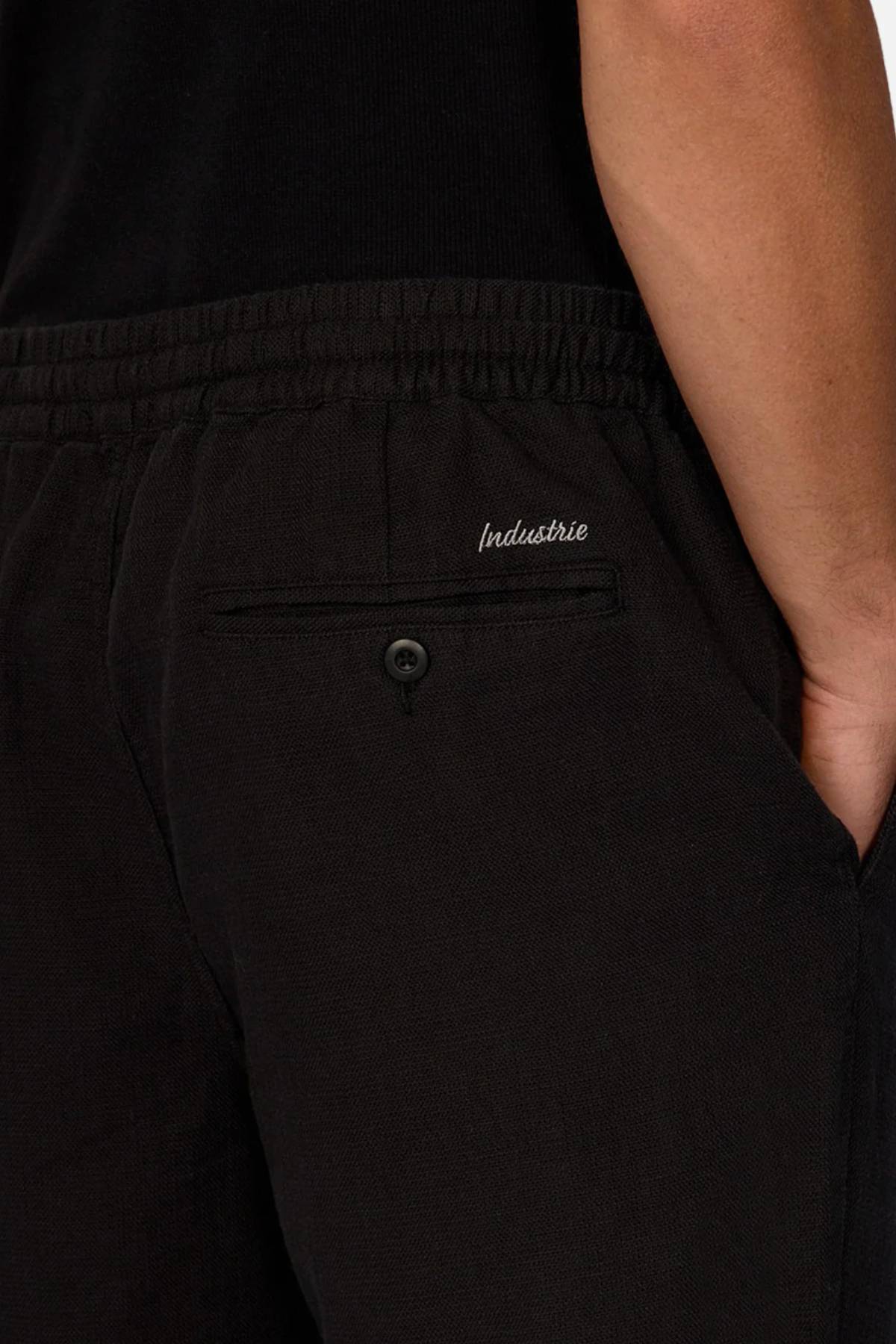 Close-up of a person wearing Industrie The Mallorca Pant in black with a hand in the pocket. Made from 100% cotton breathable fabric, the pants feature a buttoned back pocket with "Industrie" embroidery and a black top.