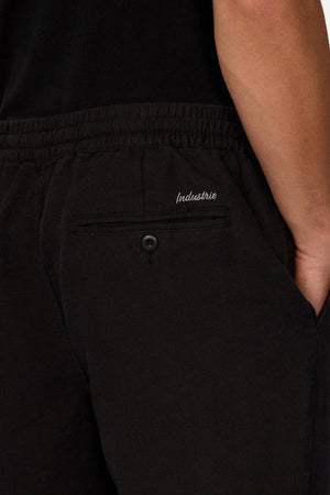 Close-up of a person wearing Industrie The Mallorca Pant in black with a hand in the pocket. Made from 100% cotton breathable fabric, the pants feature a buttoned back pocket with "Industrie" embroidery and a black top.