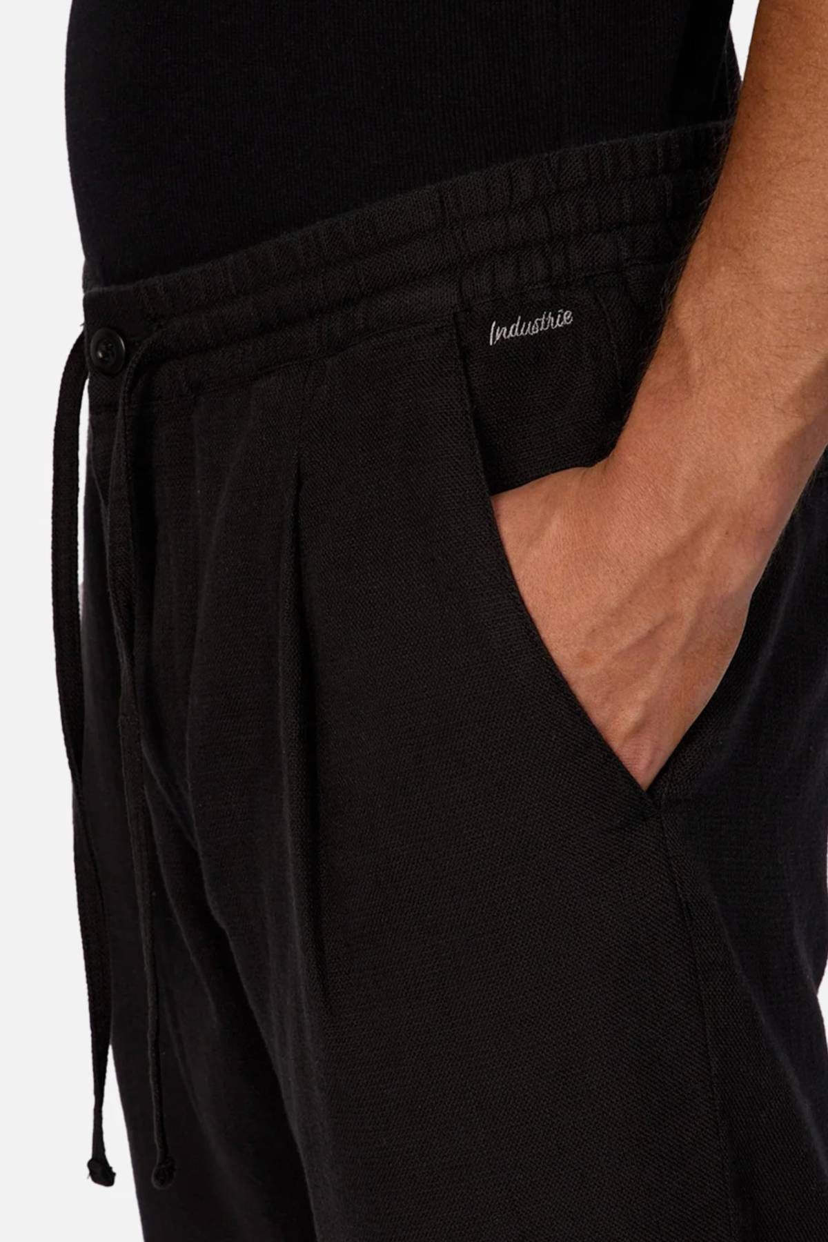 A person wears Industrie The Mallorca Pant in black, made from 100% cotton breathable fabric with a small "Industrie" logo near the pocket and one hand in the pocket, paired with a black top.