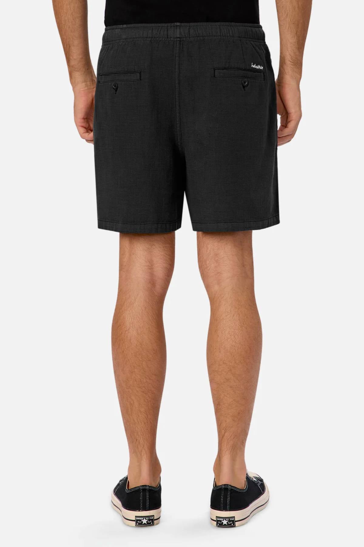 A man faces away, wearing Industrie The Mallorca Short in black cotton with back pockets, paired with a black shirt and black canvas sneakers with white soles, set against a plain light background.